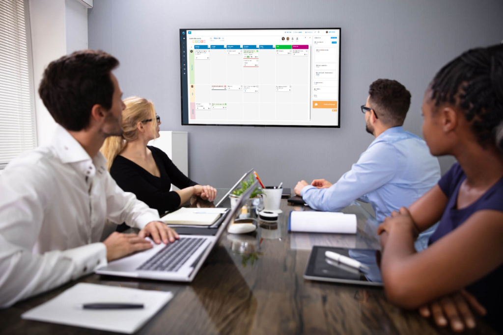 A four member team gathered in a meeting room with DigiLEAN on a wall monitor